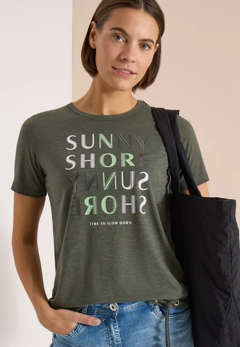 Person in olivgrünem T-Shirt mit gedrucktem 'SUNNY SHORE' Textdesign und schwarzer Tasche über der Schulter.